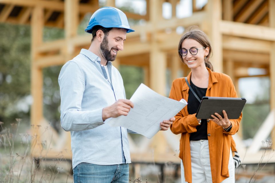 Builder with a female client near the wooden house structure on the construction site outdoors. Building and designing wooden frame house concept
