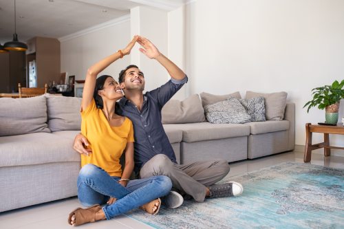 Handsome young man with beautiful indian woman dreaming a new home. Smiling married couple moves to new apartment with copy space. Happy middle eastern couple making roof with hands symbol of new home